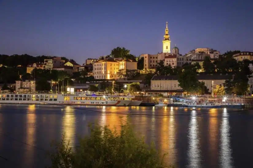Belgrade city skyline at night with the illuminated St. Michael's Cathedral spire and boats on the Sava River