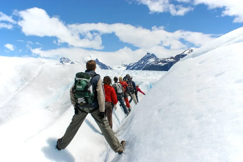 A group of hikers traverses a snowy mountain trail under a clear blue sky.