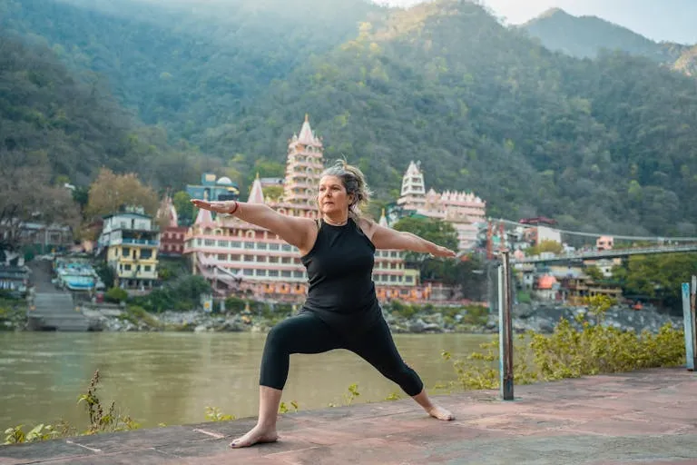 A senior woman doing yoga by the river with Rishikesh's iconic temple in the background.