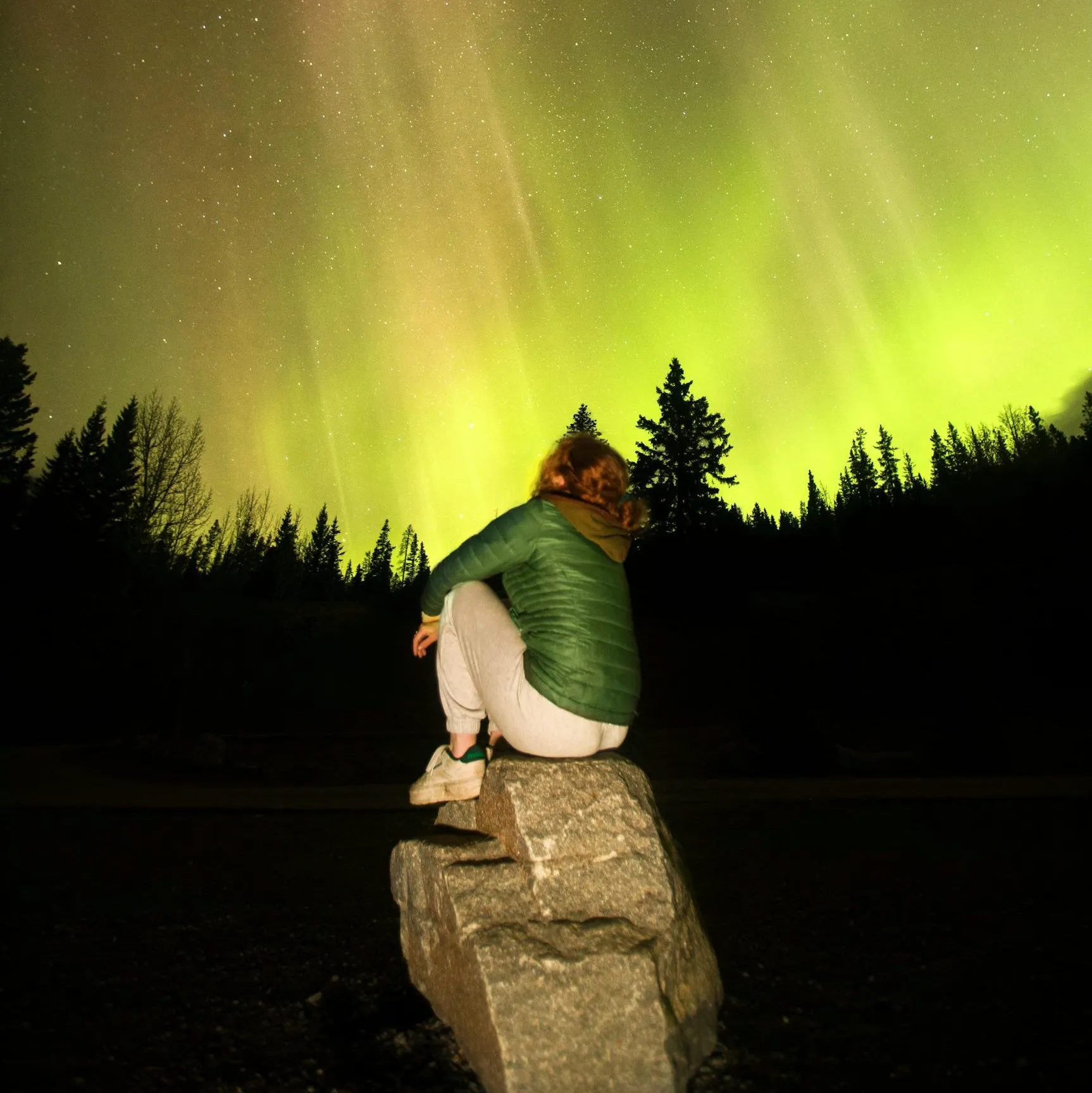 A woman sits on a rock, admiring the aurora borealis in the Canadian night sky.