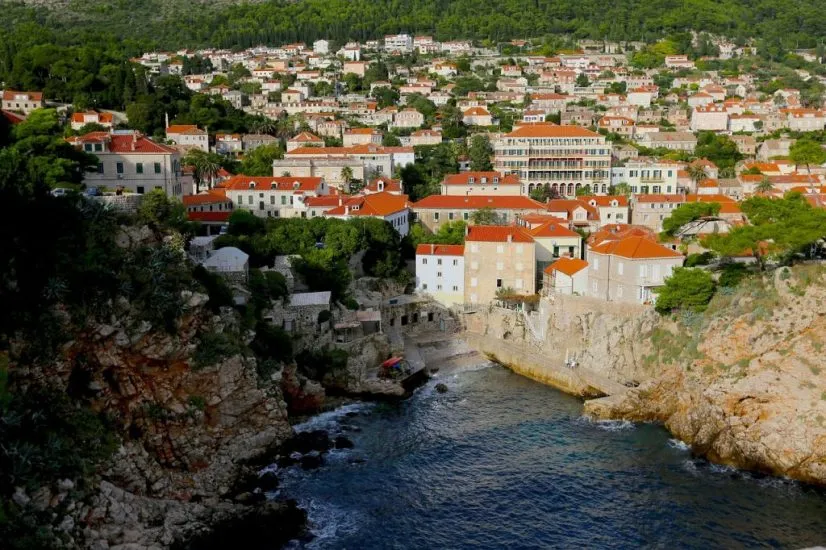 Red-roofed buildings of Dubrovnik clustered on a hillside overlooking a small rocky cove and blue sea