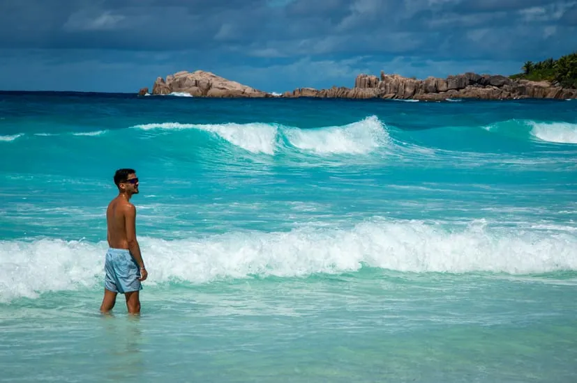 Man standing in the turquoise waters of Anse Source d'Argent, Seychelles, under clear summer skies.