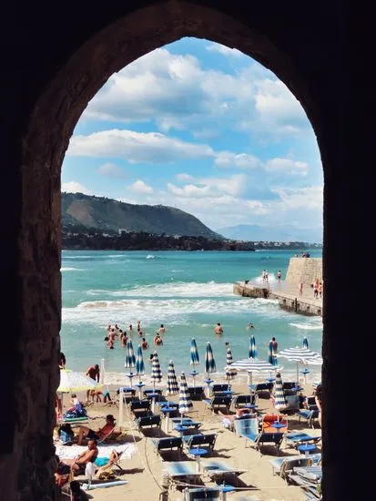 Picturesque view of Cefalù beach through an arch, capturing vacationers and the serene sea.
