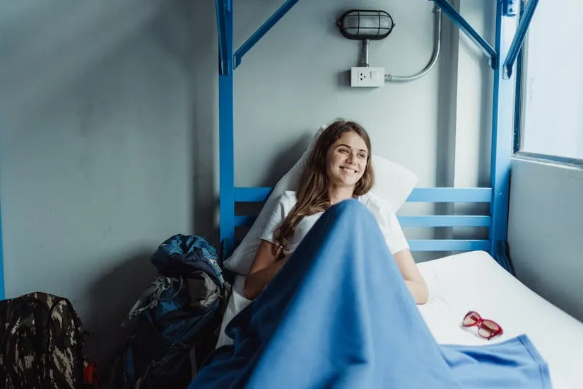 A smiling woman relaxes in a hostel bunk bed with her travel backpacks nearby