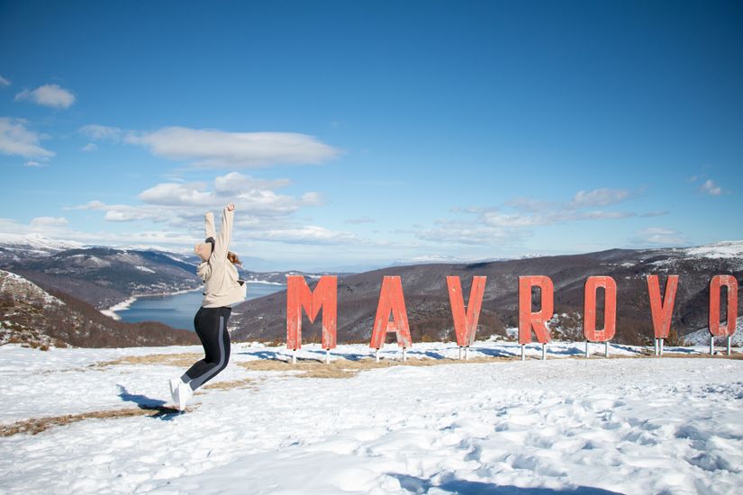 Woman jumping cheerfully in the snow beside Mavrovo sign in North Macedonia.